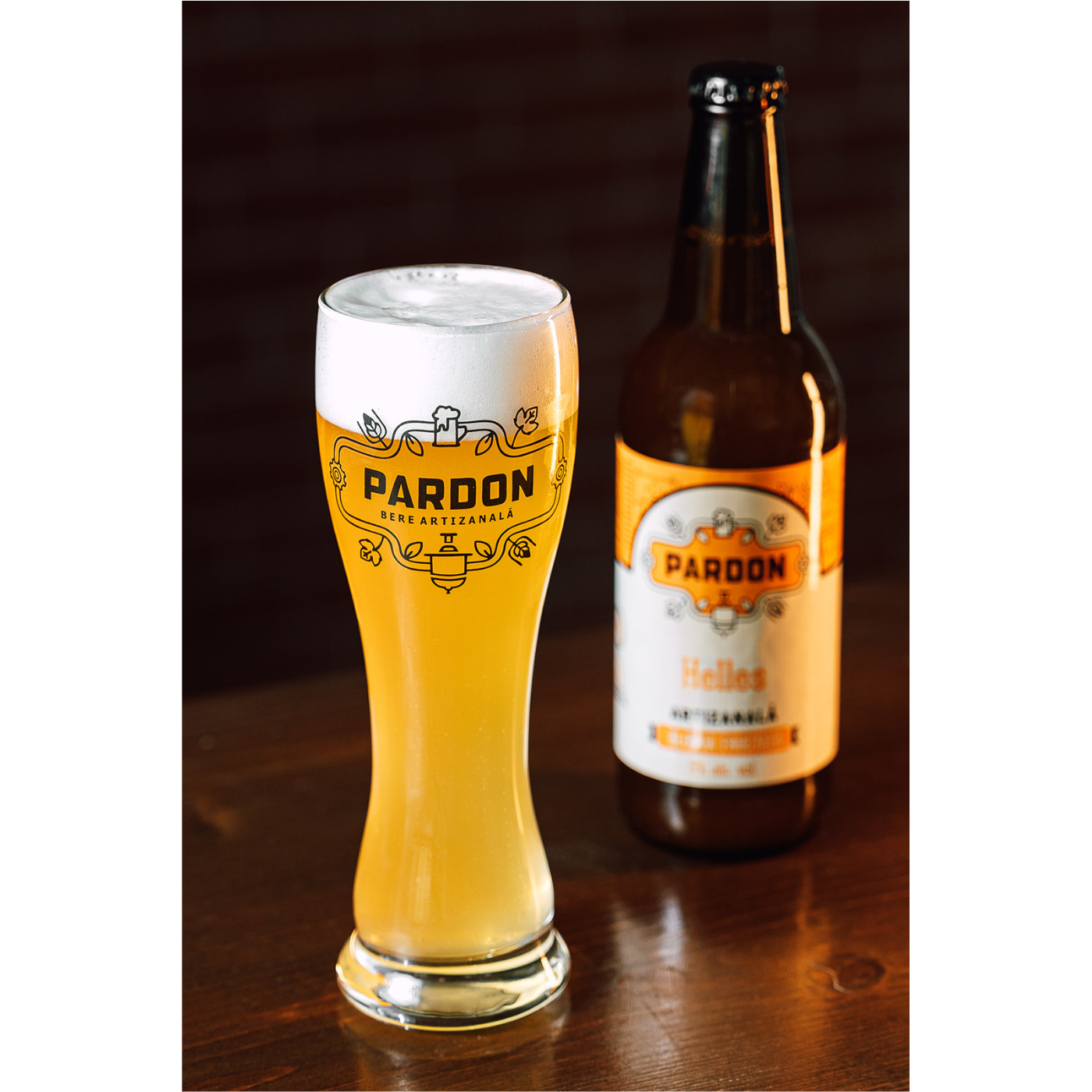 Pahar cu bere artizanala nefiltrata Pardon Helles asezat pe masă lângă sticla de 500ml