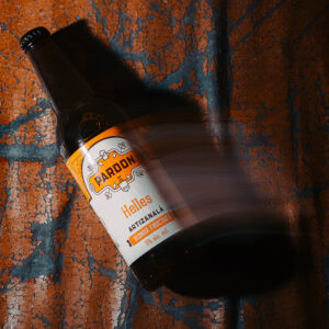 Sticlă de bere nefiltrată Pardon Helles artizanală, blondă fructată cu 5% alcool, fotografiată pe fundal artistic texturat