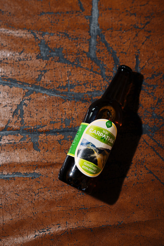 Sticlă de bere Pardon Carpathia artizanală, blondă ecologică 5% alcool, fotografiată pe fundal artistic texturat